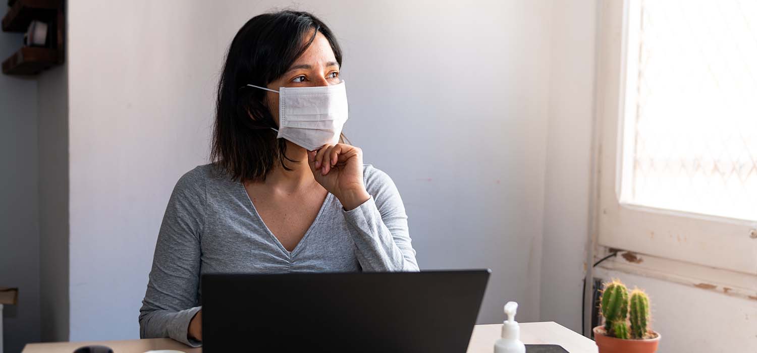 Woman wearing mask with a laptop in front of her, looking out of window